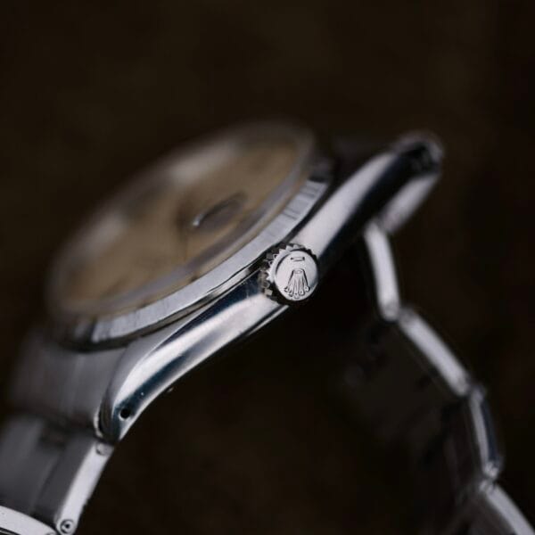 Close-up of a silver Rolex Oyster Perpetual Datejust 6605 Cal. 1066 wristwatch, highlighting the crown with part of the watch face visible. The timepiece has a metallic band set against a dark, blurred background.