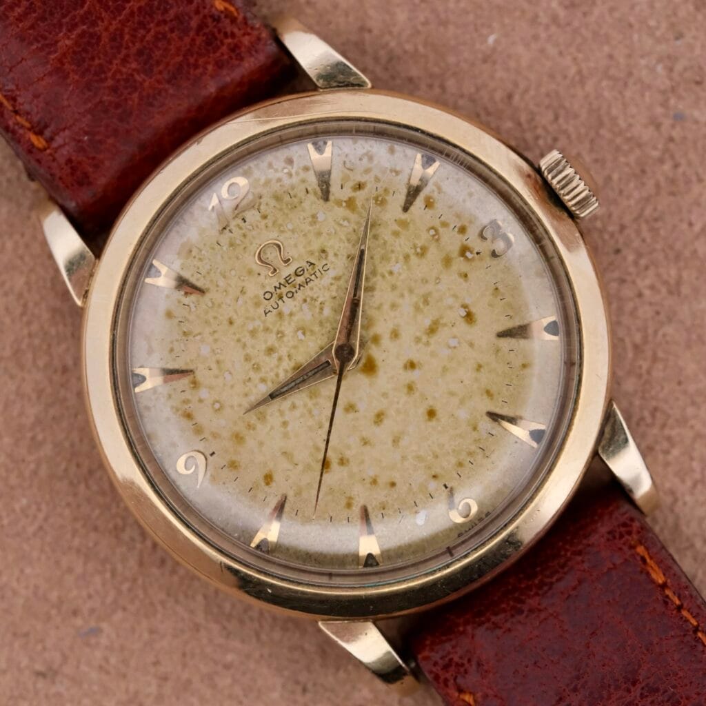 Close-up of an Omega Automatic Spotted Patina Cal. 355 wristwatch with a cream speckled dial, gold indices, and brown leather strap. The watch hands and Omega logo are prominently visible on the dial.