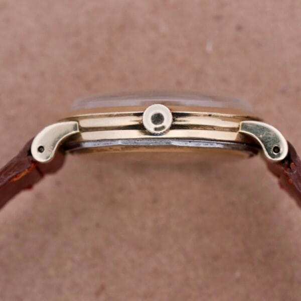 Side view of an Omega Automatic Spotted Patina Cal. 355 gold wristwatch with a round case and brown leather strap, highlighting the crown and lugs. Elegantly placed against a matching brown background.