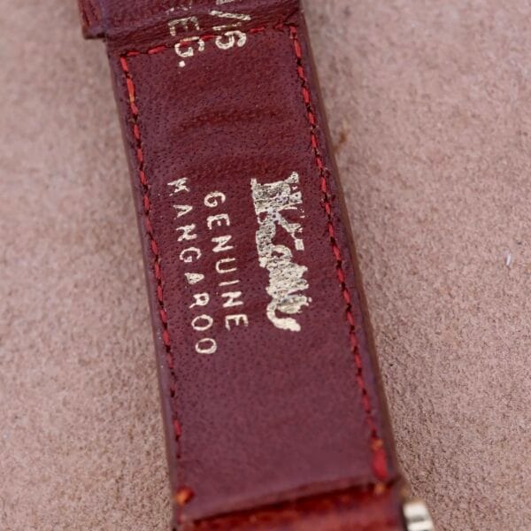 Close-up of a brown leather strap with GENUINE KANGAROO stamped in gold, showcasing Omega Automatic Spotted Patina Cal. 355s craftsmanship. Visible stitching and slight wear are present against a textured beige background.