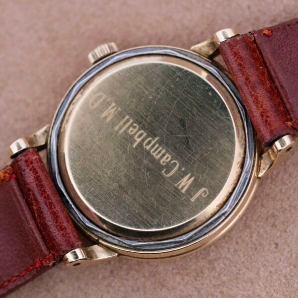 Close-up of the back of an Omega Automatic Spotted Patina Cal. 355 wristwatch featuring a brown leather strap. The vintage casing is engraved J W Campbell M.D. and shows small scratches and wear, enhancing its timeless charm.