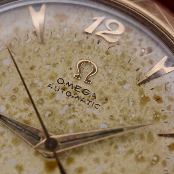 Close-up of an Omega Automatic Spotted Patina Cal. 355 watch face, featuring a speckled patina. The dial highlights gold hour markers and hands, with the iconic Omega logo and OMEGA AUTOMATIC text prominently displayed.