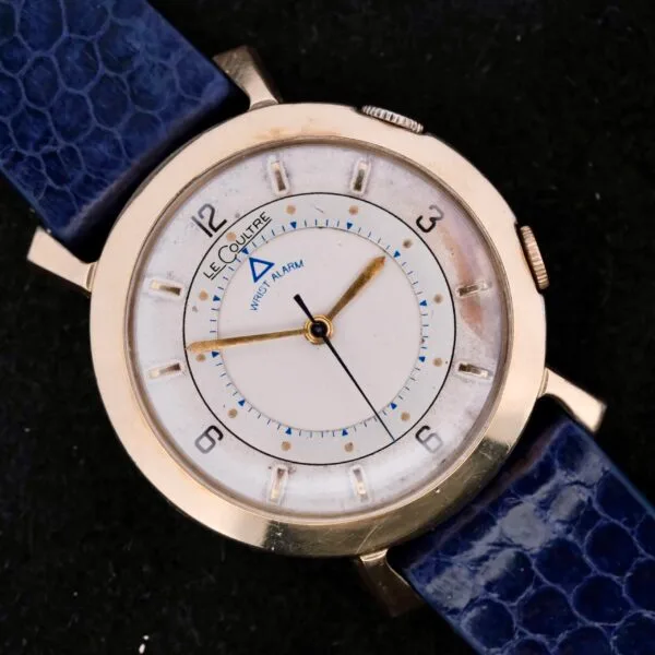 The vintage Lecoultre Wrist Alarm watch has a gold-tone case, white dial, numeral hour markers, and a blue leather strap. It features two crowns for its wrist alarm function and displays LeCoultre at the top.