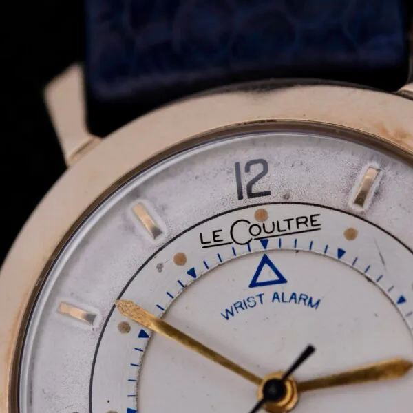 A close-up of the vintage Lecoultre Wrist Alarm reveals a white face with the brand name, gold hour markers and hands, a blue triangle for the alarm, all set on a striking blue leather strap.