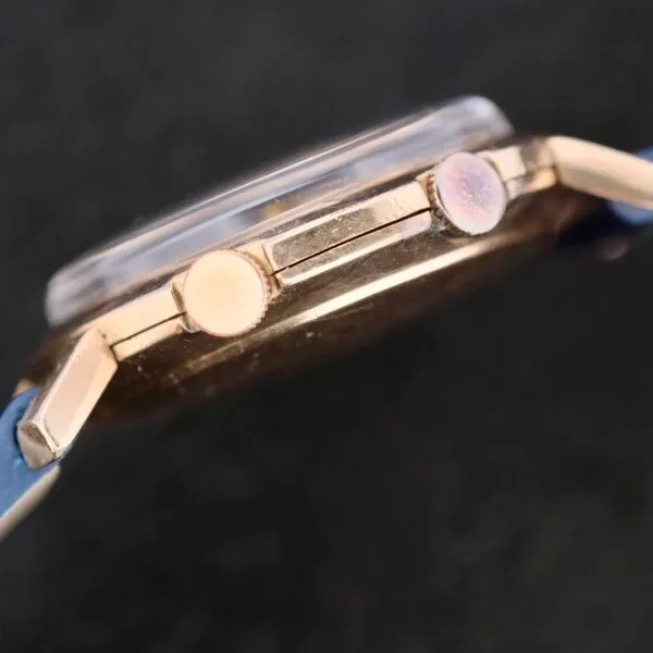 Close-up of a vintage Lecoultre Wrist Alarm watch showcasing a round crown and button on a textured black background. The watch face, known for its wrist alarm feature, is slightly visible with an elegant blue strap.