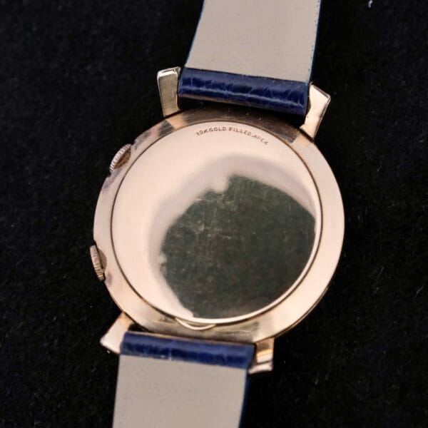 The image displays the Lecoultre Wrist Alarm, highlighting a 10K gold-filled case back and a cream and blue strap with slight surface scratches, set against a dark background.