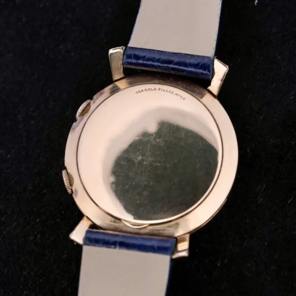 The image displays the Lecoultre Wrist Alarm, highlighting a 10K gold-filled case back and a cream and blue strap with slight surface scratches, set against a dark background.