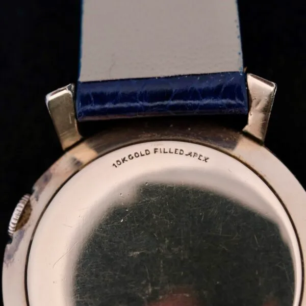 Close-up of a watch back labeled 10K Gold Filled Apex. This Lecoultre Wrist Alarm has a light strap with a dark blue section near the case, adding elegance despite surface scratches and wear.