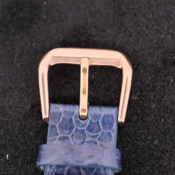 Close-up of a Lecoultre Wrist Alarms gold buckle on a blue textured leather strap against a black background. The shiny buckle enhances the straps intricate pattern, reflecting the watchs sophisticated design.
