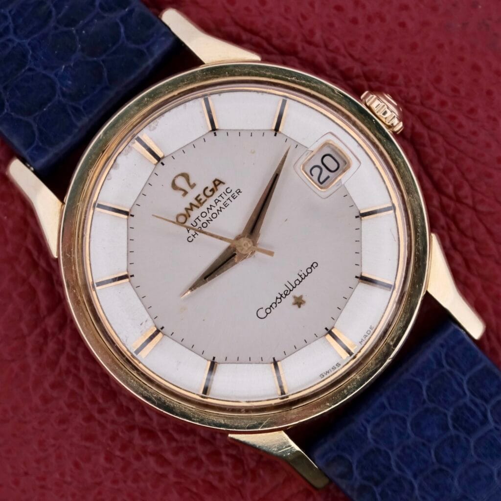 The Omega Constellation Pie-Pan Automatic 561 features a vintage round gold case, white dial with Automatic Chronometer inscribed, date window at 3 oclock, gold hour markers and hands, complemented by a dark blue leather strap against a striking red background.