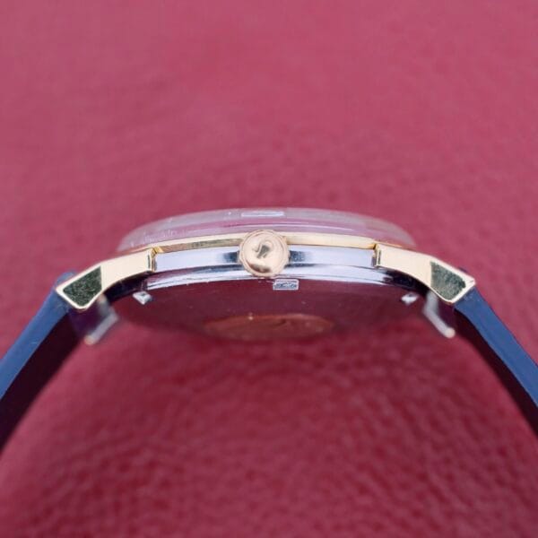 Side view of the Omega Constellation Pie-Pan Automatic 561 with a blue strap and gold-toned crown against a textured red background. The metallic case gleams elegantly, and the crown features a small logo.