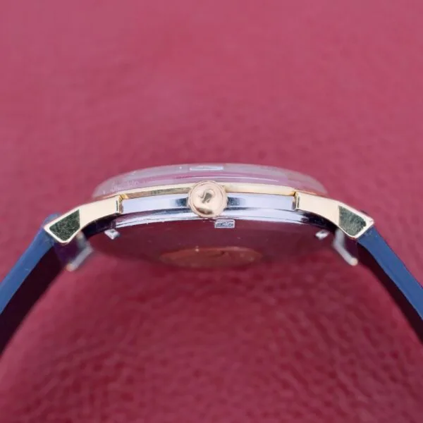 Side view of the Omega Constellation Pie-Pan Automatic 561 with a blue strap and gold-toned crown against a textured red background. The metallic case gleams elegantly, and the crown features a small logo.