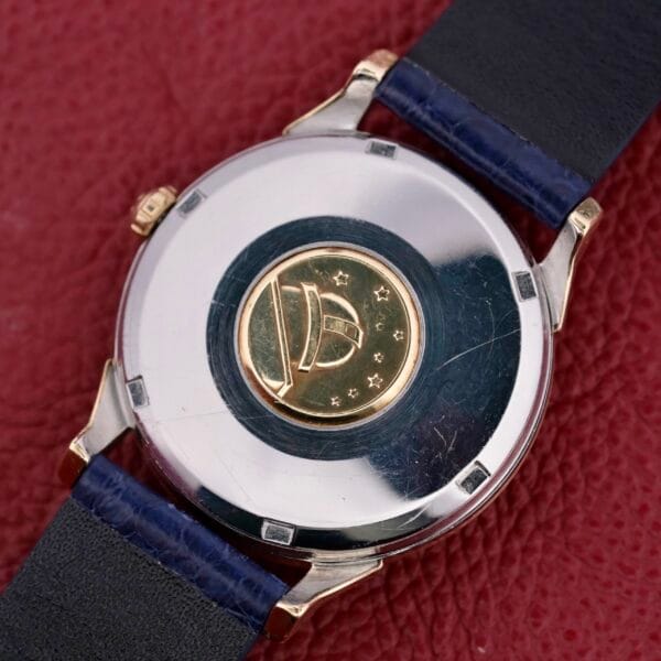 The Omega Constellation Pie-Pan Automatic 561 features a circular gold medallion on the back with a dome and star design set against a textured red background, complemented by an elegant black leather strap.