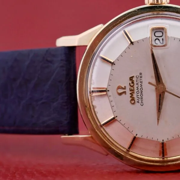 Close-up of a vintage Omega Constellation Pie-Pan Automatic 561 watch with a gold case, white dial, and date display at 3 oclock. The timepiece features a dark blue leather strap set elegantly against a muted red background.