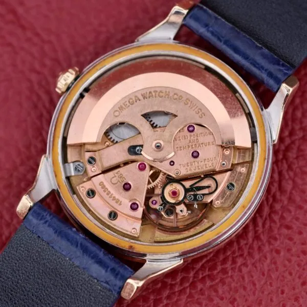 Close-up of the Omega Constellation Pie-Pan Automatic 561s intricate inner mechanism, highlighting detailed gears with visible engravings. The pinkish-gold finish contrasts with a dark blue strap against a textured red background.