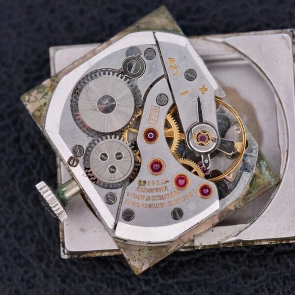 A close-up reveals the intricate mechanism of the Longines Tank 14k White Gold Diamond Dial Cal. 370, showcasing visible gears, screws, and jewels against a black backdrop. The metallic components are compactly arranged in its rectangular Tank formation, highlighting the engineering marvel.