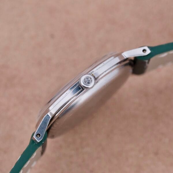 Side view of a minimalist Hamilton Thin-O-Matic Linen Dial T-502 Micro-Rotor wristwatch featuring a silver case and green leather strap. The crown, adorned with a sleek logo, is set against a plain brown surface.