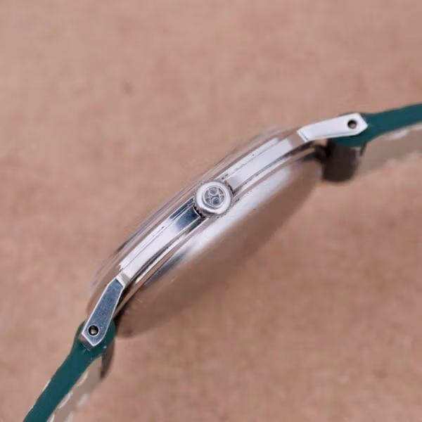 Side view of a minimalist Hamilton Thin-O-Matic Linen Dial T-502 Micro-Rotor wristwatch featuring a silver case and green leather strap. The crown, adorned with a sleek logo, is set against a plain brown surface.