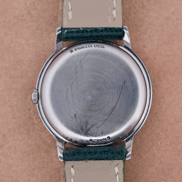 The image showcases a Hamilton Thin-O-Matic Linen Dial T-502 Micro-Rotor. The stainless steel case back displays visible scratches, while the leather strap with contrasting stitching enhances its classic look. Its round design features a crown on the left side, embodying timeless elegance.