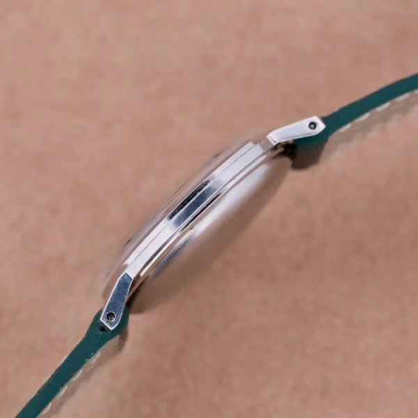 A close-up side view of the Hamilton Thin-O-Matic Linen Dial T-502 Micro-Rotor wristwatch highlights its green leather strap, with a sleek polished silver case and delicate lugs against a textured beige background, showcasing timeless elegance.