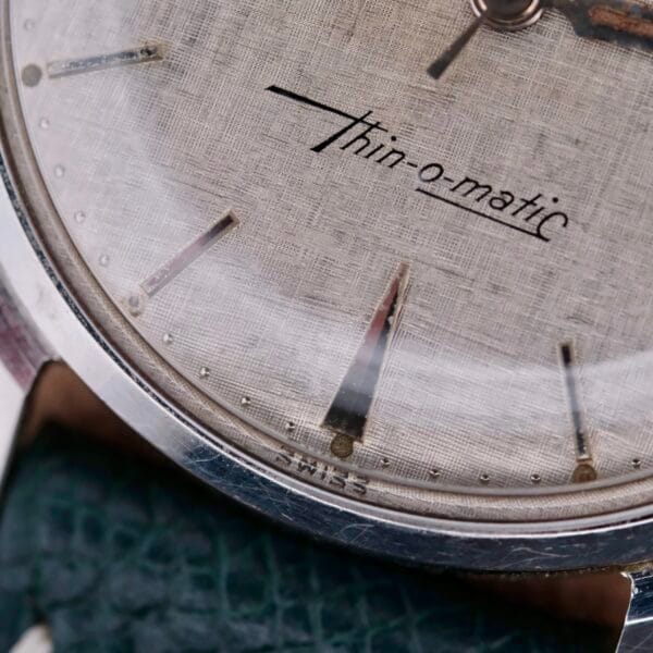 Close-up of a vintage Hamilton Thin-O-Matic Linen Dial T-502 Micro-Rotor watch face, featuring thin-o-matic lettering. The textured dial with minimalist markers is encased in sleek silver, accented by a green textured strap and the word Swiss at its base.