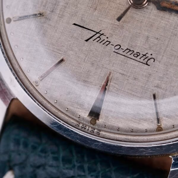 Close-up of a vintage Hamilton Thin-O-Matic Linen Dial T-502 Micro-Rotor watch face, featuring thin-o-matic lettering. The textured dial with minimalist markers is encased in sleek silver, accented by a green textured strap and the word Swiss at its base.