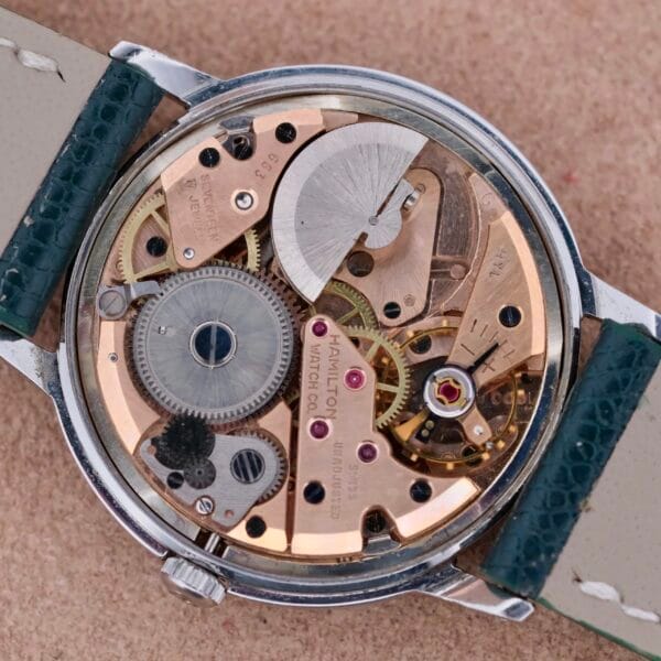 The image displays the intricate movement of a Hamilton Thin-O-Matic Linen Dial T-502 Micro-Rotor watch, showcasing exposed gears, jewels, and metal components with the back case removed. The brand name is engraved on the mechanics, and it comes with a leather strap.