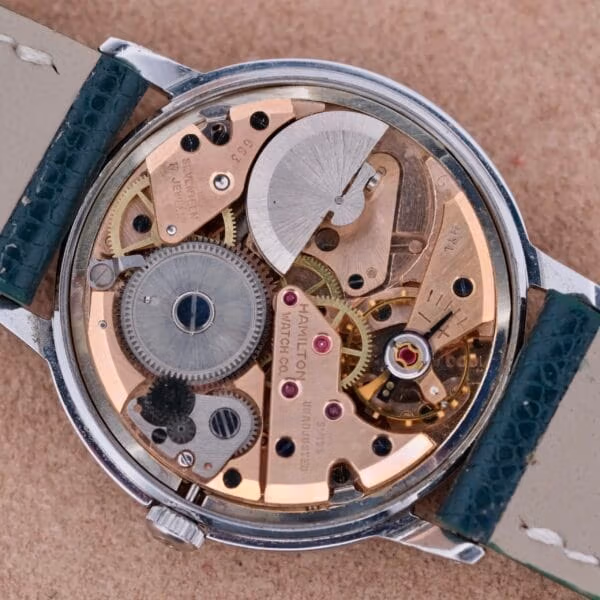 The image displays the intricate movement of a Hamilton Thin-O-Matic Linen Dial T-502 Micro-Rotor watch, showcasing exposed gears, jewels, and metal components with the back case removed. The brand name is engraved on the mechanics, and it comes with a leather strap.