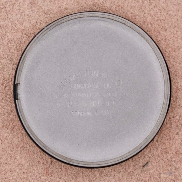 A round, silver lid inscribed with HAMILTON MFG CO LANCASTER, PA STAINLESS STEEL PAT. PEND. STAR M. C. CO. elegantly rests on a light brown surface, echoing the sophistication of the Hamilton Thin-O-Matic Linen Dial T-502 Micro-Rotor.