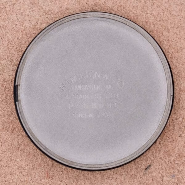A round, silver lid inscribed with HAMILTON MFG CO LANCASTER, PA STAINLESS STEEL PAT. PEND. STAR M. C. CO. elegantly rests on a light brown surface, echoing the sophistication of the Hamilton Thin-O-Matic Linen Dial T-502 Micro-Rotor.