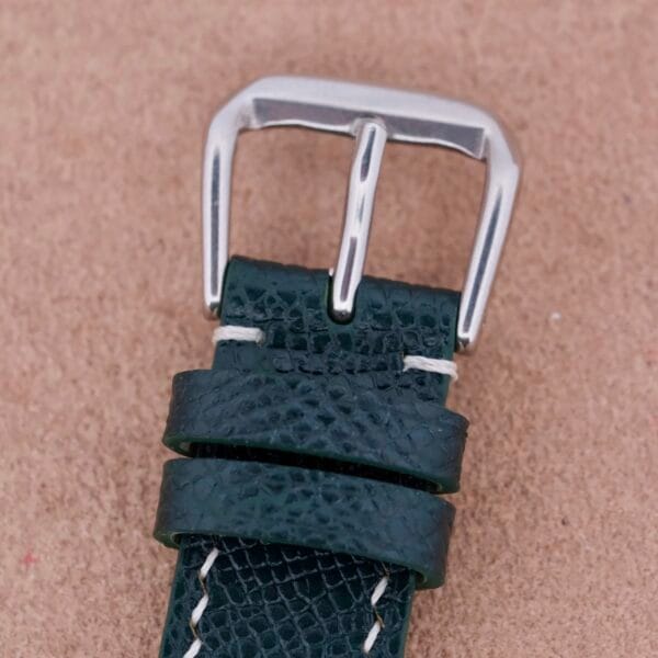 Close-up of a green leather watch strap with a silver buckle, inspired by the classic Hamilton Thin-O-Matic Linen Dial T-502 Micro-Rotor. Featuring two loops and white stitching, it contrasts beautifully against a neutral beige background.