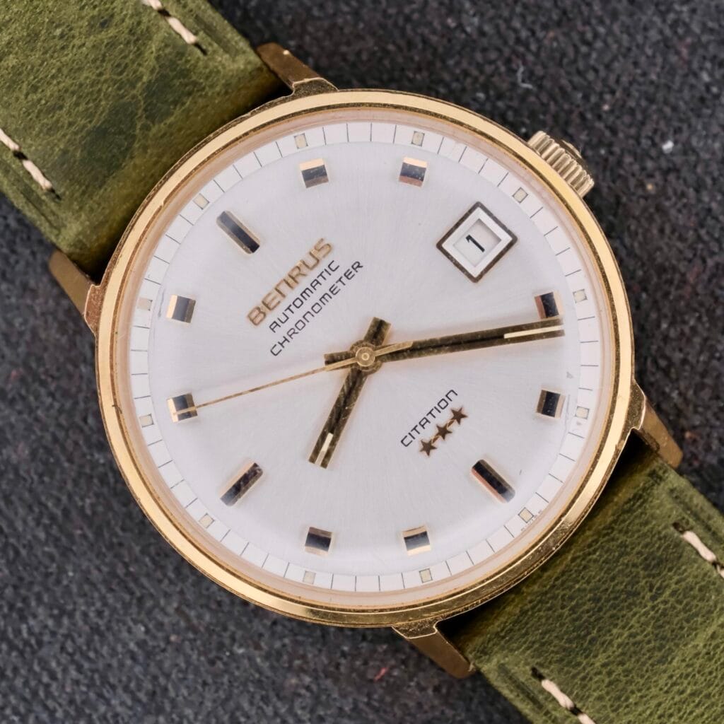 The vintage Benrus Citation Automatic Chronometer Date watch showcases a white face and gold casing with Automatic Chronometer text. It includes three hands, a date display, and is elegantly completed with a green leather strap featuring white stitching.