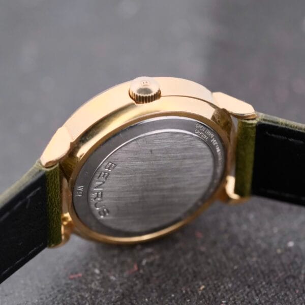 A side view close-up of a Benrus Citation Automatic Chronometer Date wristwatch, highlighting its gold casing and black leather strap. The silver back panel is engraved with BENRUS. It rests elegantly on a dark, textured surface.