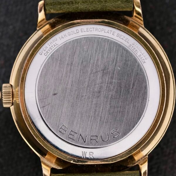 The Benrus Citation Automatic Chronometer Date wristwatch features a metal case back engraved with BENRUS and Open Thru 14K Gold Electroplate Bezel Crystal, as well as Stainless Steel Back. Complemented by a green strap, it offers an elegant design.