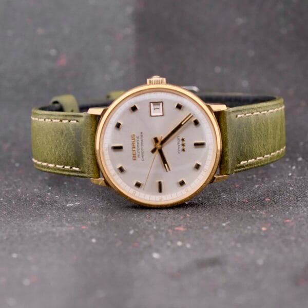 The Benrus Citation Automatic Chronometer Date watch, with a green leather strap and gold-colored casing, rests on a speckled gray surface. It features a silver dial, black hour markers, and a date window at the top, exemplifying classic elegance.