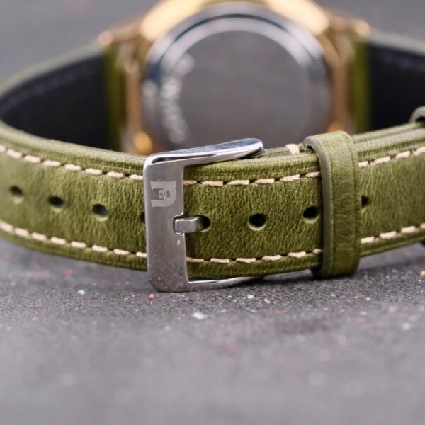 Close-up of a Benrus Citation Automatic Chronometer Date with an olive green leather strap and a silver buckle engraved with a small logo, set against a gray textured background with subtle colorful specks, showcasing the watchs elegance.