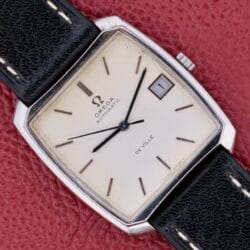 An Omega Deville Automatic with a tonneau case and black leather strap rests elegantly on a red textured surface.