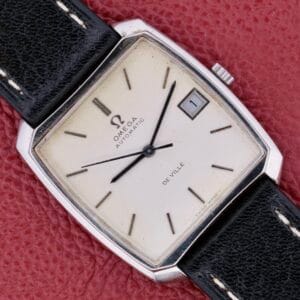 An Omega Deville Automatic with a tonneau case and black leather strap rests elegantly on a red textured surface.