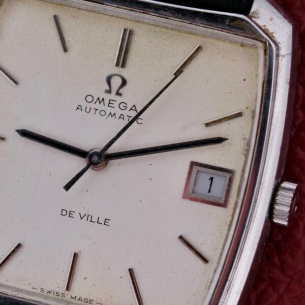 A close-up of the Omega Deville Automatic Tonneau Case wristwatch reveals its square face, featuring a cream hue and minimalist hour markers and hands. The date window displays 1, while Swiss Made highlights the watchs precision and craftsmanship.