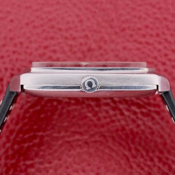 Close-up of an Omega Deville Automatic Tonneau Case watch with a black leather strap on a textured red background. The side view features a logo-engraved crown, showcasing the watchs sleek design and craftsmanship.