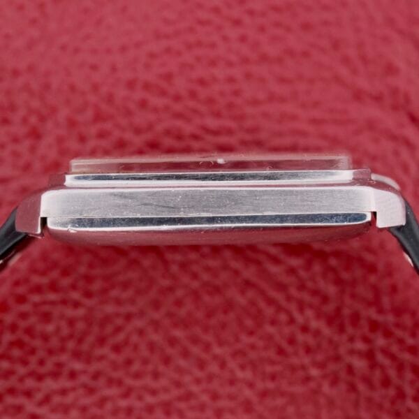 Close-up side view of an Omega Deville Automatic Tonneau Case watch, featuring a sleek silver case with a black strap, set against a textured red background.