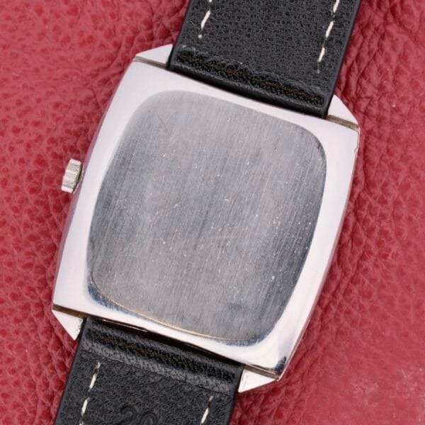 The image displays the back of an Omega Deville Automatic Tonneau Case wristwatch, with a silver finish and black leather strap, set on a textured red surface. The slightly scratched back panel tells of its journey through time.