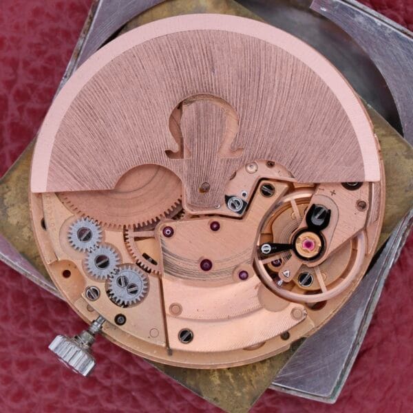 Close-up of an Omega Deville Automatic Tonneau Case watch movement showcasing intricate gears, levers, and a winding crown on a red background, emphasizing the craftsmanship and detailing of the watchs internal mechanisms.