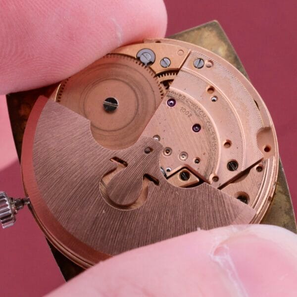 Close-up of fingers holding the intricate mechanism of an Omega Deville Automatic Tonneau Case watch, with visible components like gears and winding mechanism against a reddish background.