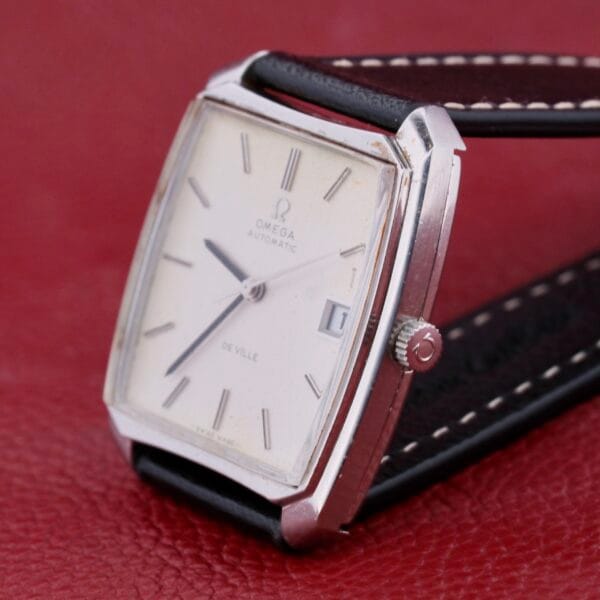 The Omega Deville Automatic Tonneau Case wristwatch features a vintage rectangular silver case, white dial, and black leather strap. It rests on red leather and includes silver hour markers, a date window, and boasts a simple, elegant design.