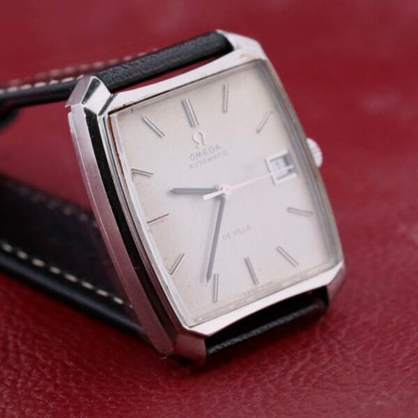The Omega Deville Automatic Tonneau Case wristwatch showcases its elegant rectangular design with a white dial, silver hour markers, silver hands, and a date window. Its sleek black leather strap complements the watch beautifully against a maroon background.