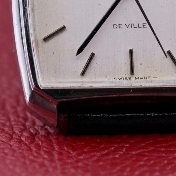 Close-up of an Omega Deville Automatic Tonneau Case watch face on a red leather surface, showcasing its minimalist design with thin, dark hour markers and hands. The words DE VILLE and SWISS MADE appear on the white dial.