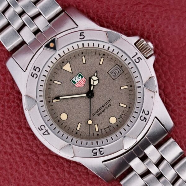 A Tag Heuer 1500 Series wristwatch with a silver metal bracelet sits on a red textured background. It features a round face, gray dial, white hour markers, and a date display at 3 oclock, blending sporty design with elegance.