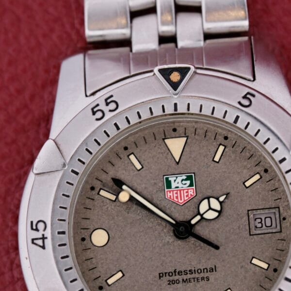 A TAG Heuer 1500 Series watch with a silver bezel lies on a red background. Its gray face showcases luminescent markers, hands, and a 3 oclock date window. The phrase professional 200 meters is elegantly above the six oclock position.