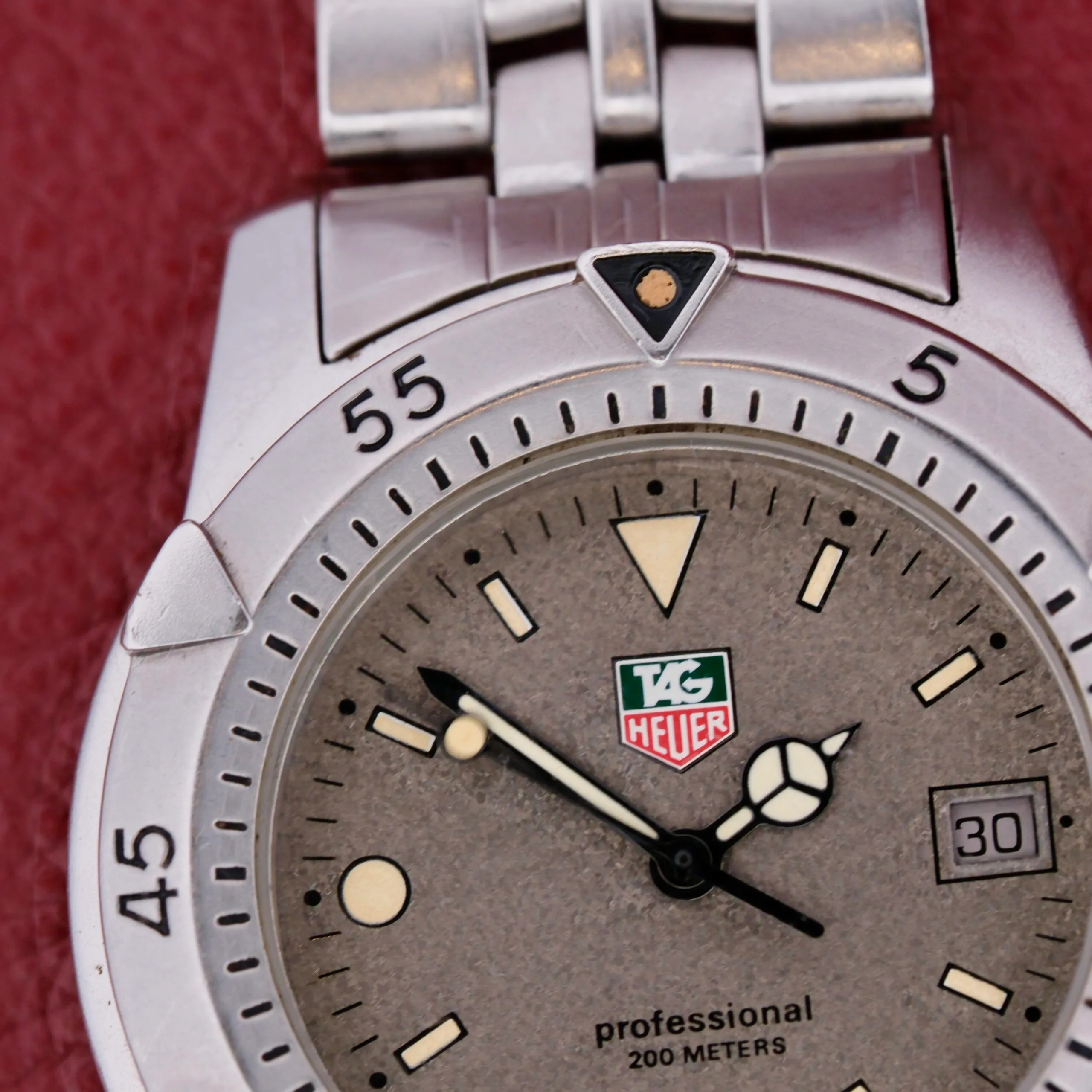 A TAG Heuer 1500 Series watch with a silver bezel lies on a red background. Its gray face showcases luminescent markers, hands, and a 3 oclock date window. The phrase professional 200 meters is elegantly above the six oclock position.